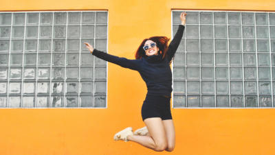 A woman in white sunglasses jumping in joy in front of a bright yellow building.